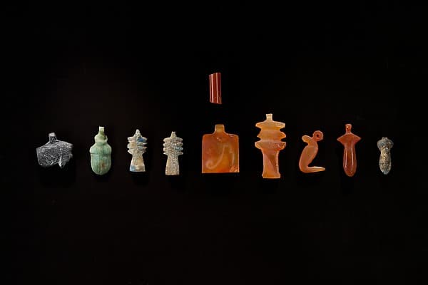 10 Amulets: 1 wedjat, 1 scarab, 3 djed pillars, 1 incised plaque, 1 uraeus, 2 wadj signs, and 1 cylindrical bead